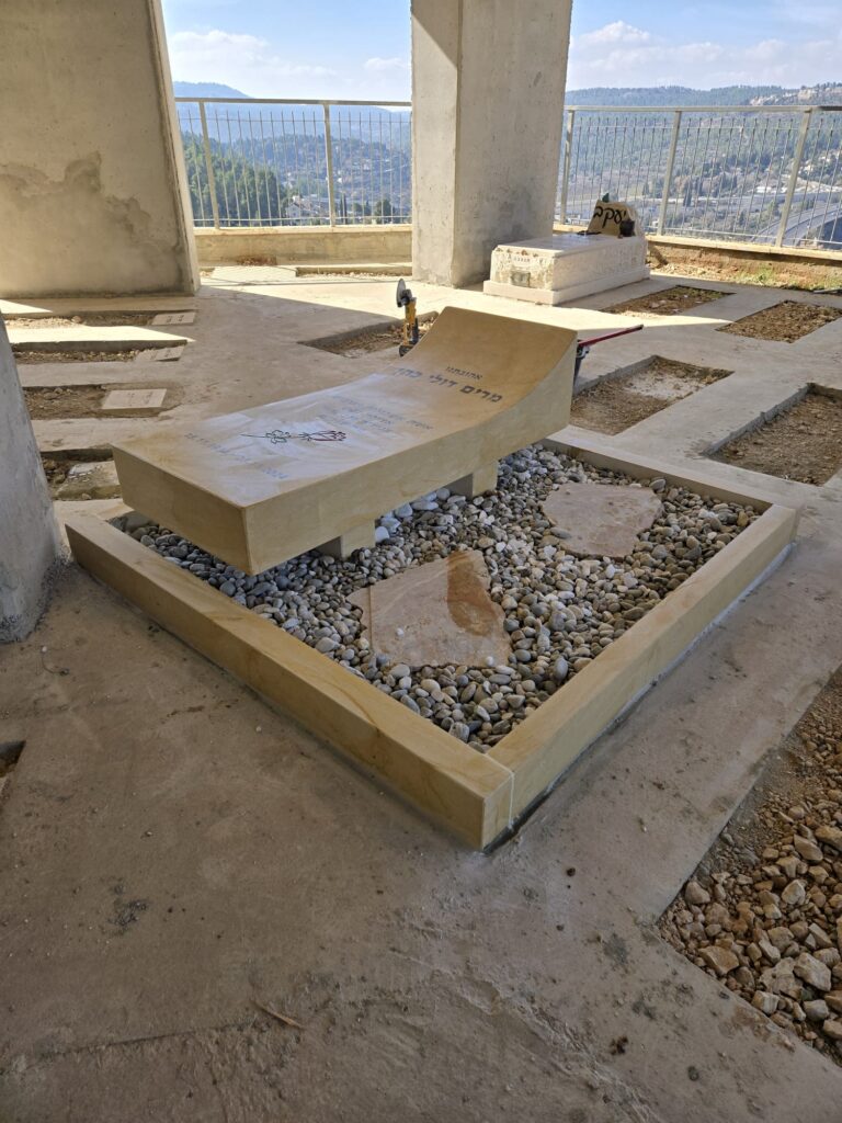 מצבה בעיצוב ייחודי עשויה שיש חברון בגוון זהוב בהיר, עם שיפוע קדמי ועדינות צורנית, מוקפת באבני נוי בירושלים.